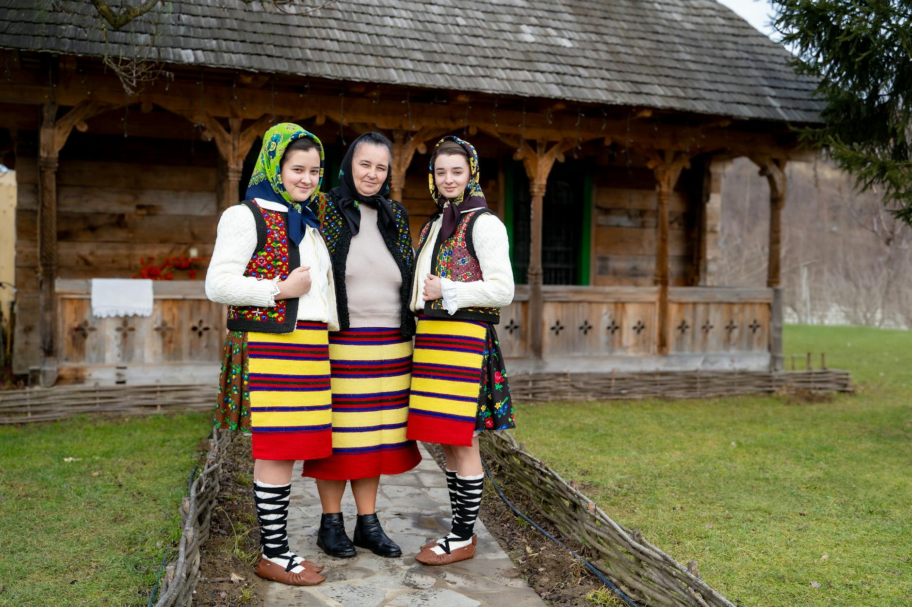 Trzy kobiety w barwnych strojach ludowych przed drewnianym domem
